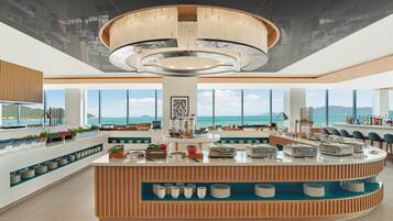 A modern hotel lobby with a central island counter, large windows with a sea view, and a high ceiling with a circular light fixture.