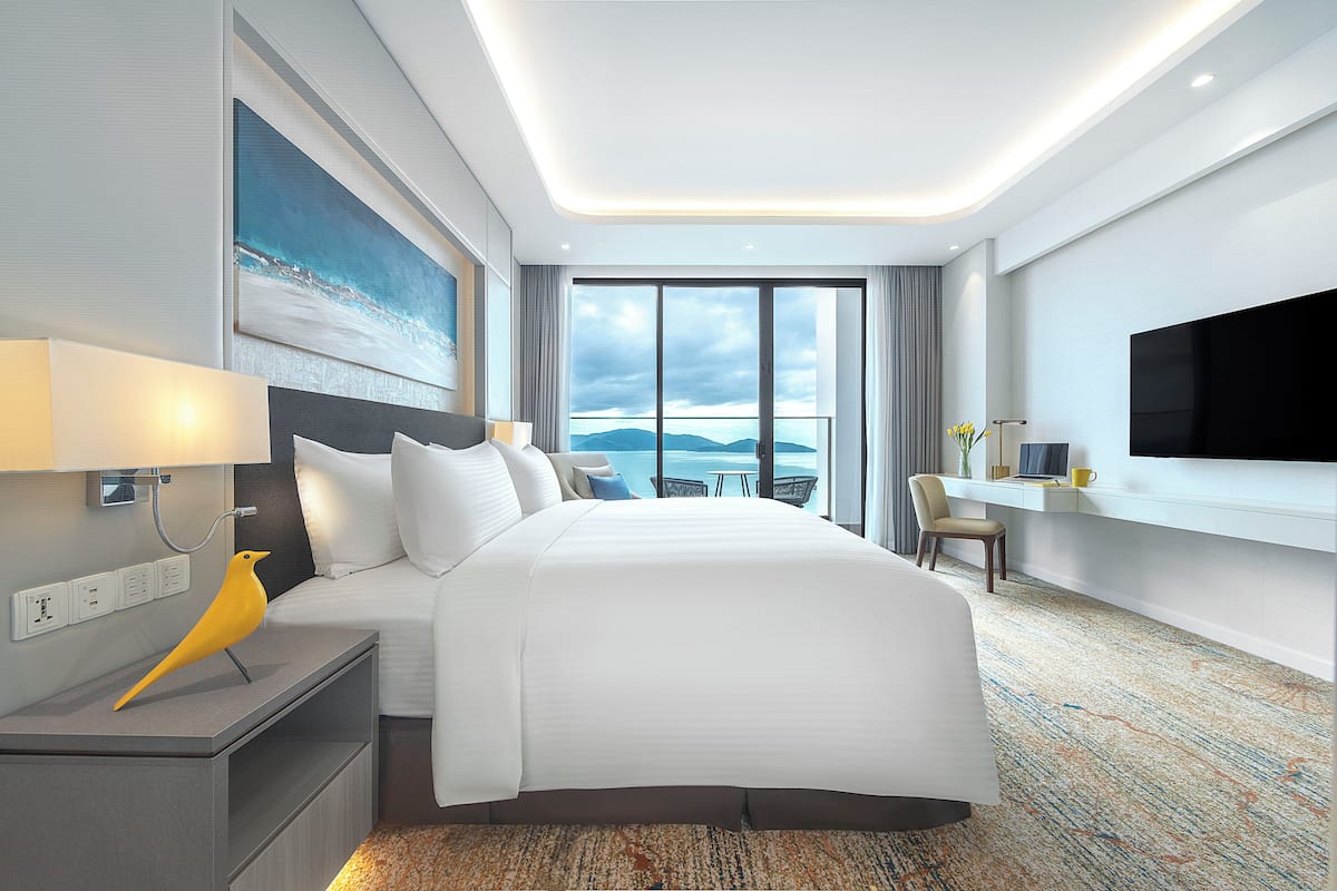 A modern hotel room with a large bed, a desk, a flat-screen TV, and a view of the sea.