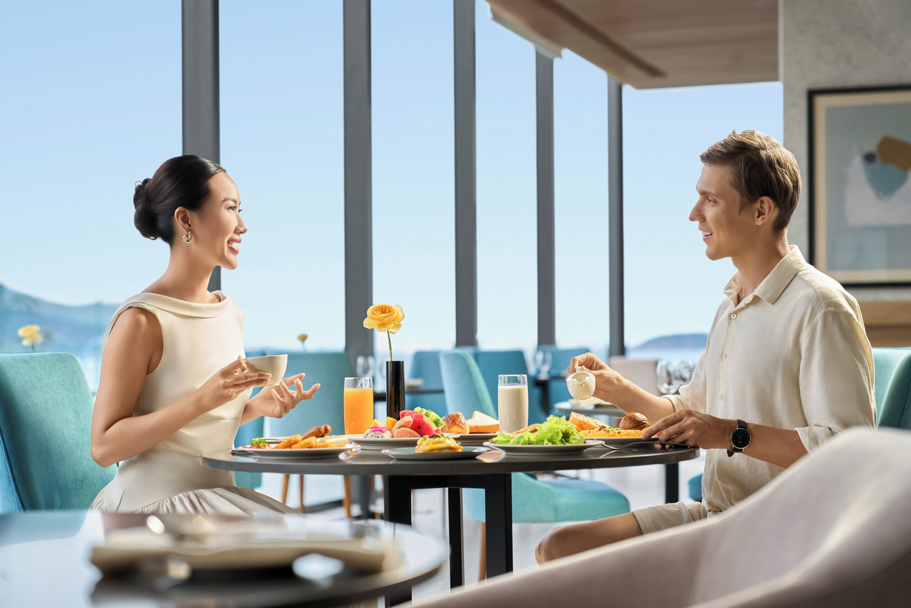 A couple dining at a table with a view of the outdoors, a vase with a flower, and various breakfast items.