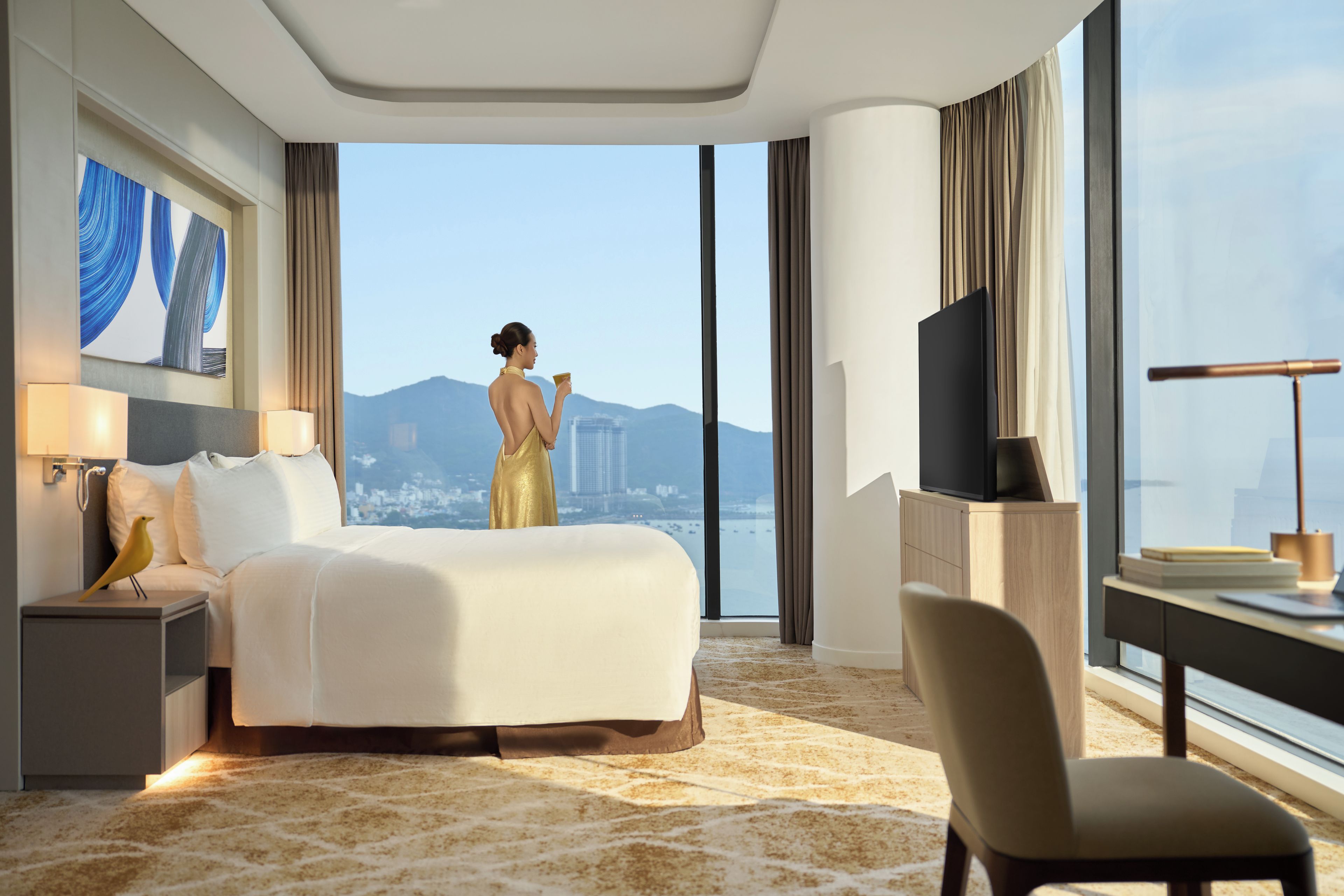A hotel room with a large bed, a desk, a chair, a TV, and a view of a cityscape.