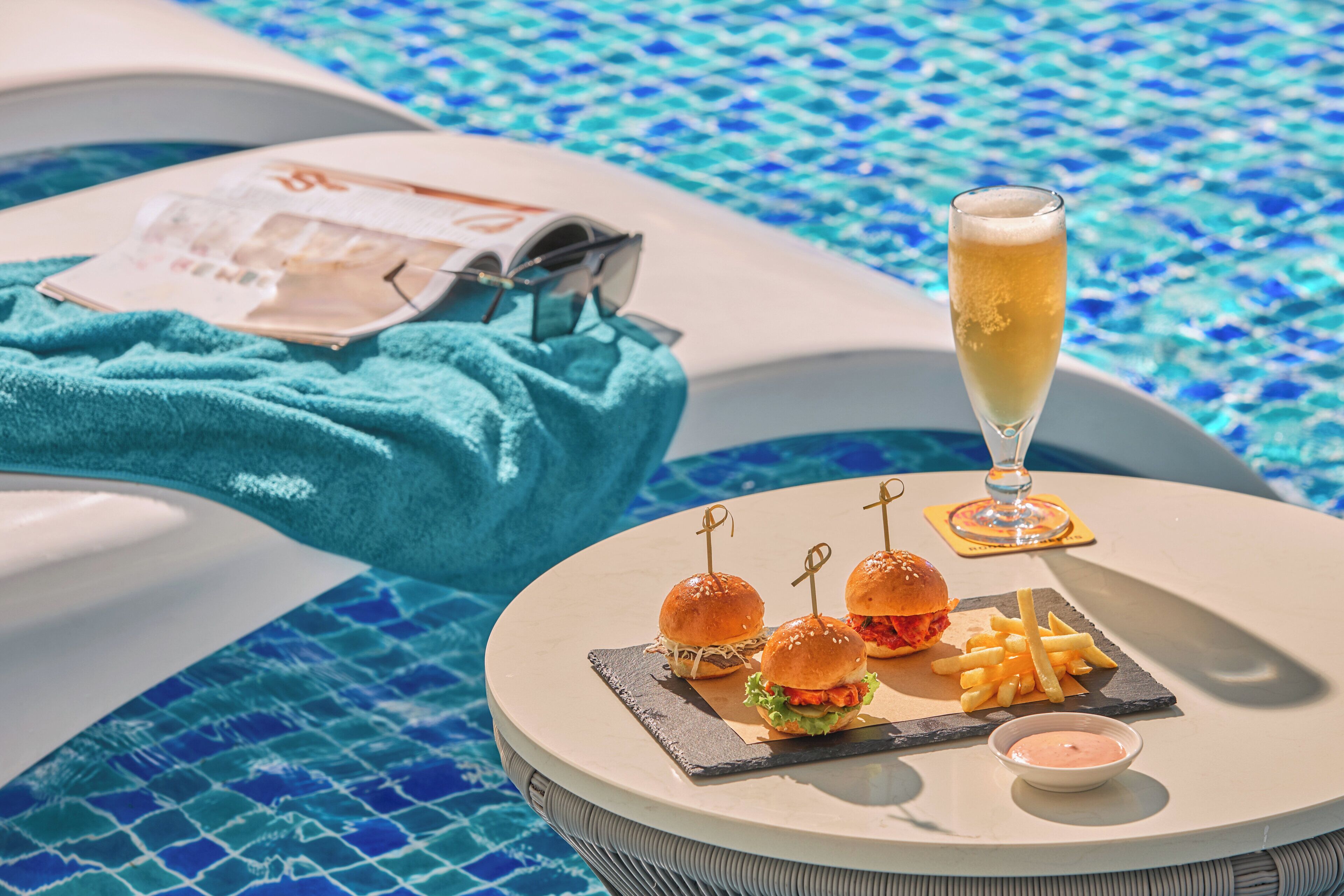 A poolside dining setup with a round table, a glass of champagne, burgers, fries, and a menu.