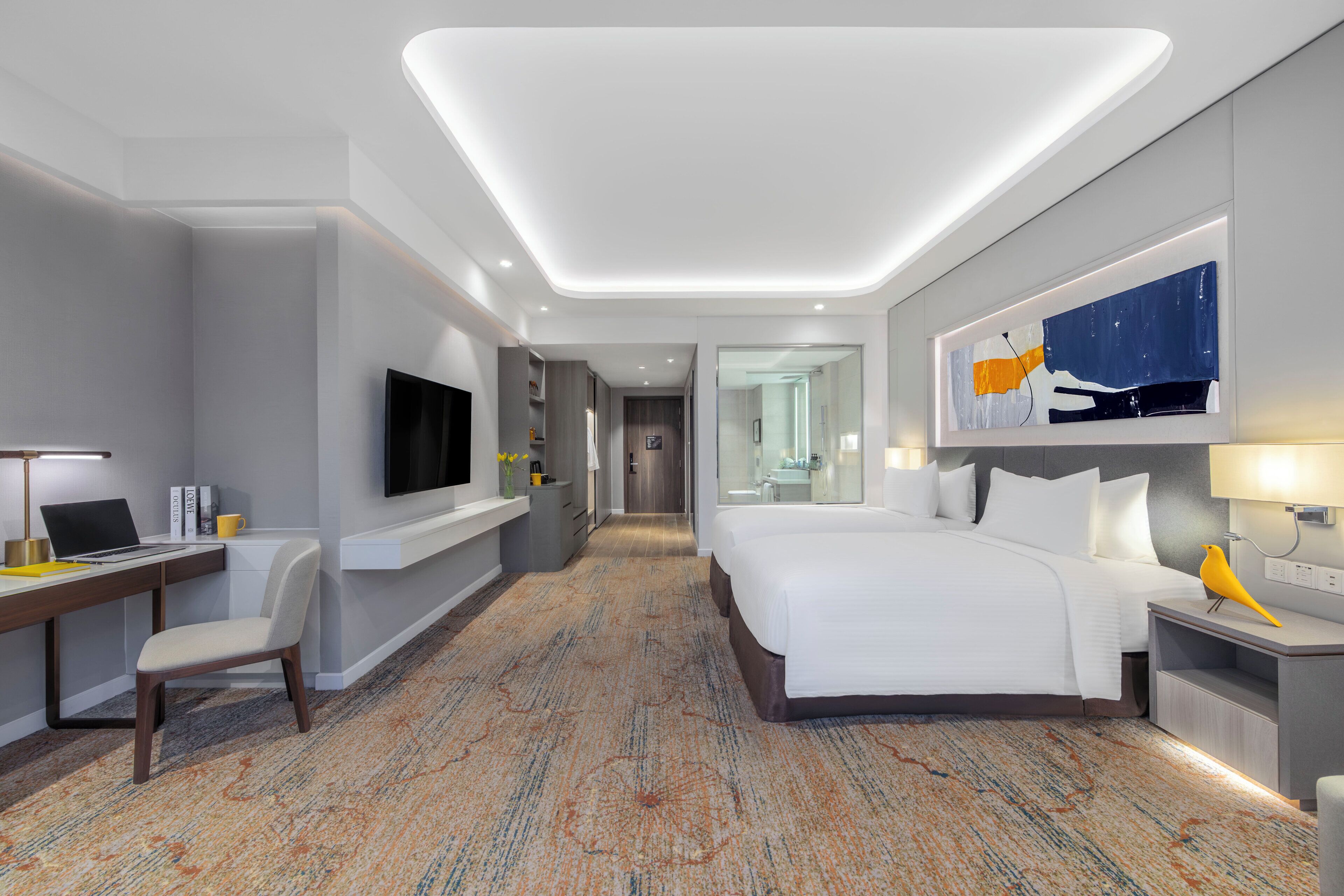 A modern hotel room with a large bed, a desk with a laptop, a flat-screen TV, and a small table with a lamp.