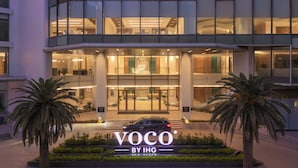 Un edificio moderno con una entrada destacada, palmeras y un cartel que dice 'VOCO BY IHG'.
