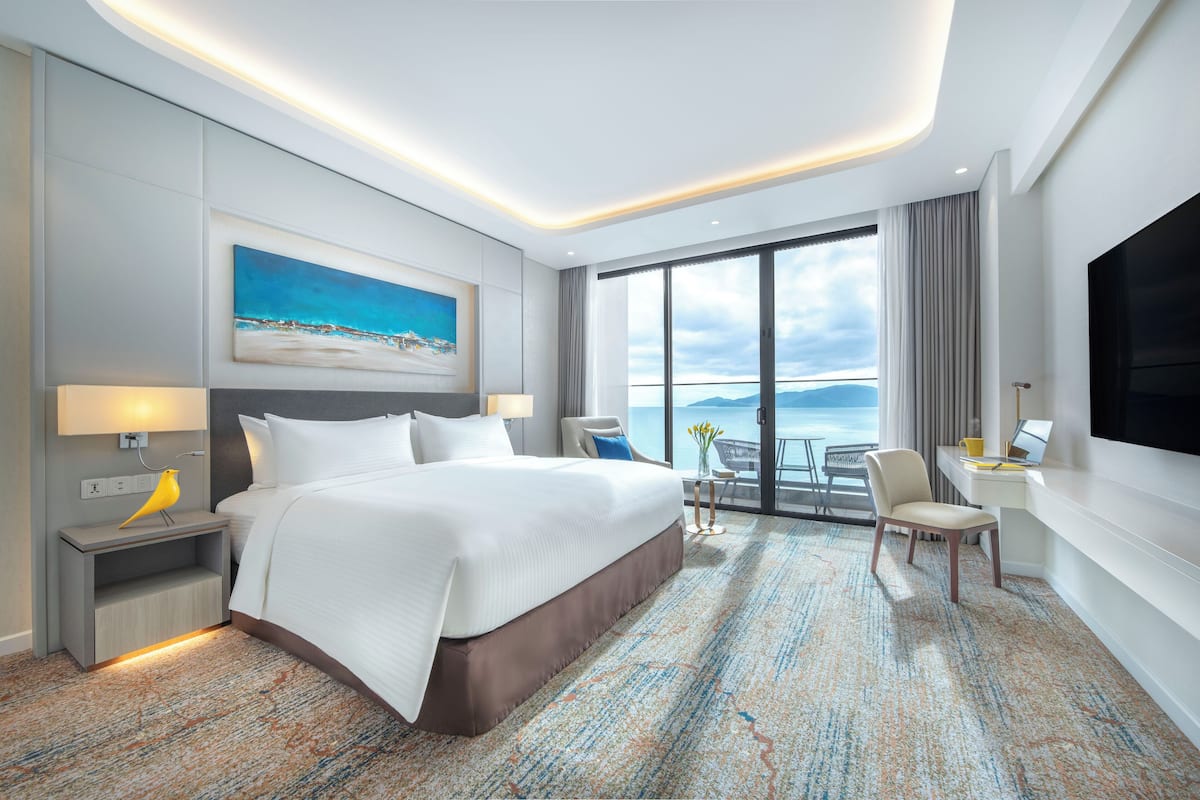A modern hotel room with a large bed, a desk, a chair, a TV, and a balcony with a view of the sea.