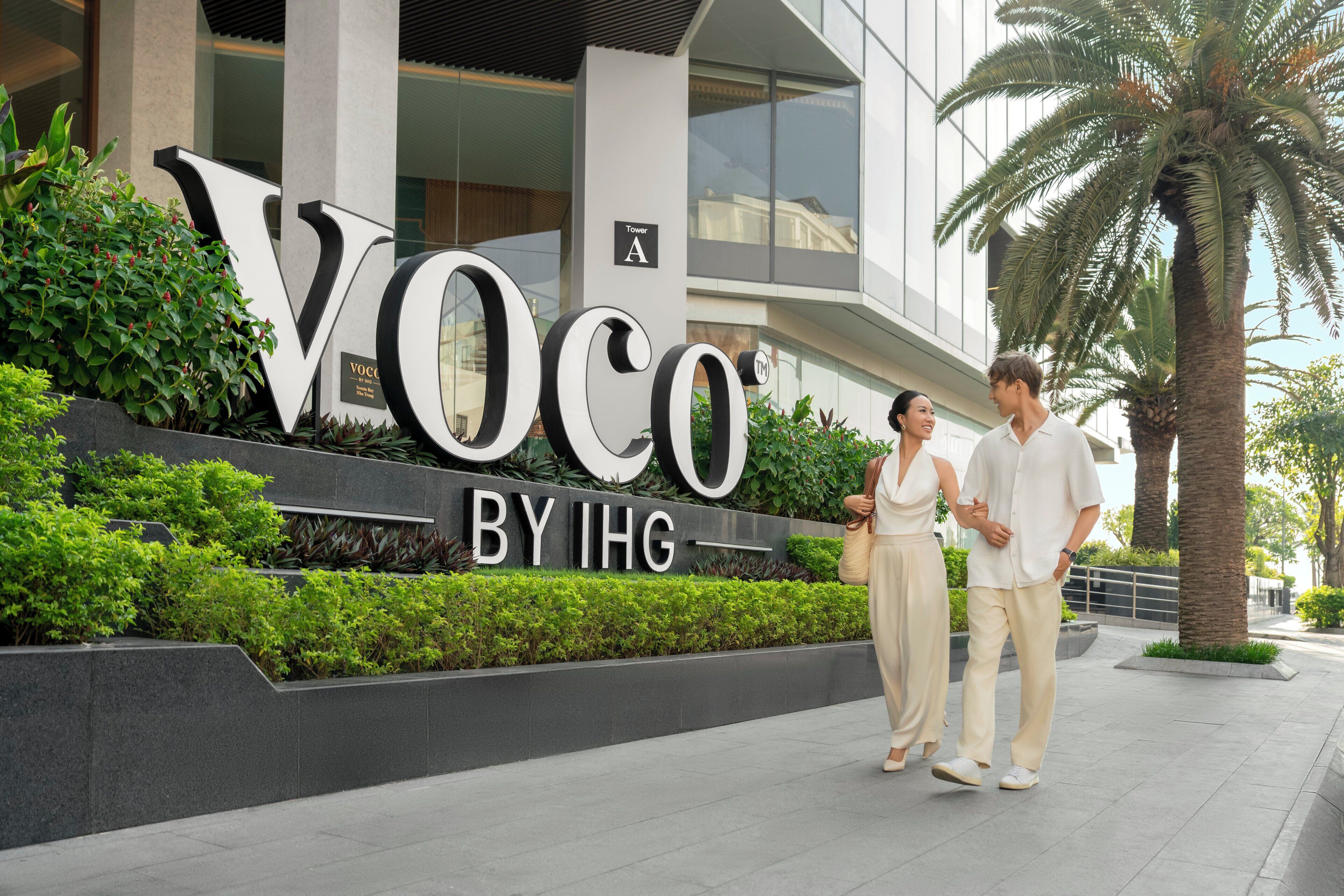 A modern building with a large sign reading 'Voco by IHG' and a landscaped area with palm trees.