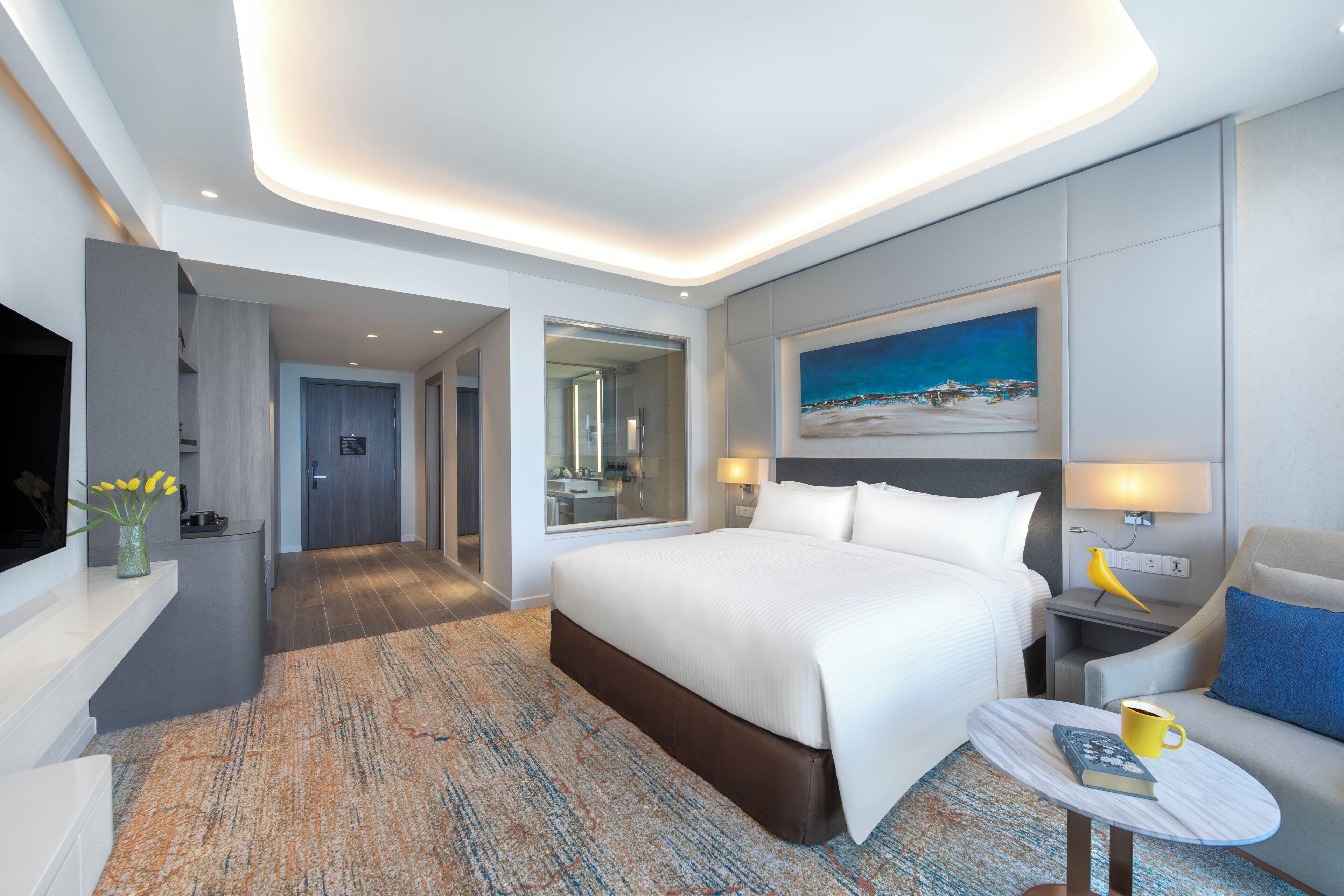 A modern hotel room with a large bed, a small table with a book and a mug, a nightstand with a lamp, a painting on the wall, and a bathroom visible through an open door.