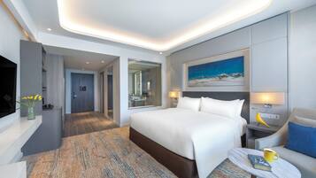 A modern hotel room with a large bed, a small table with a book and a mug, a nightstand with a lamp, a painting on the wall, and a bathroom visible through an open door.