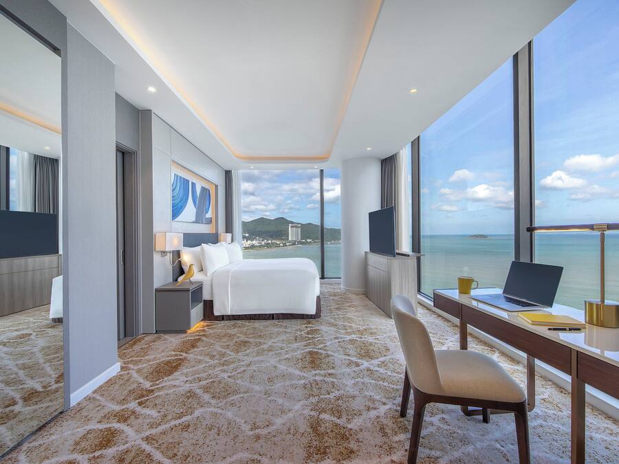 A modern hotel room with a large bed, a desk with a laptop, and a view of the sea.