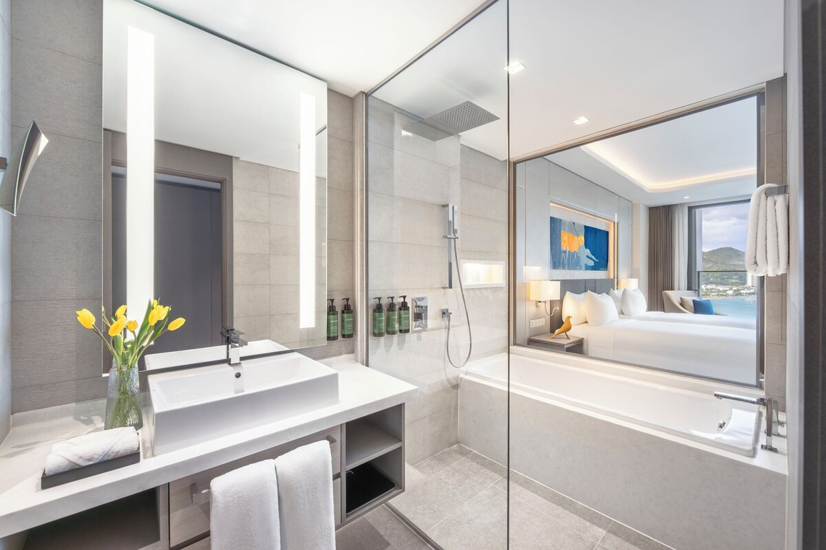A modern hotel bathroom with a large mirror, a white sink, a glass-enclosed shower, and a view of a bedroom with a bed and a painting.