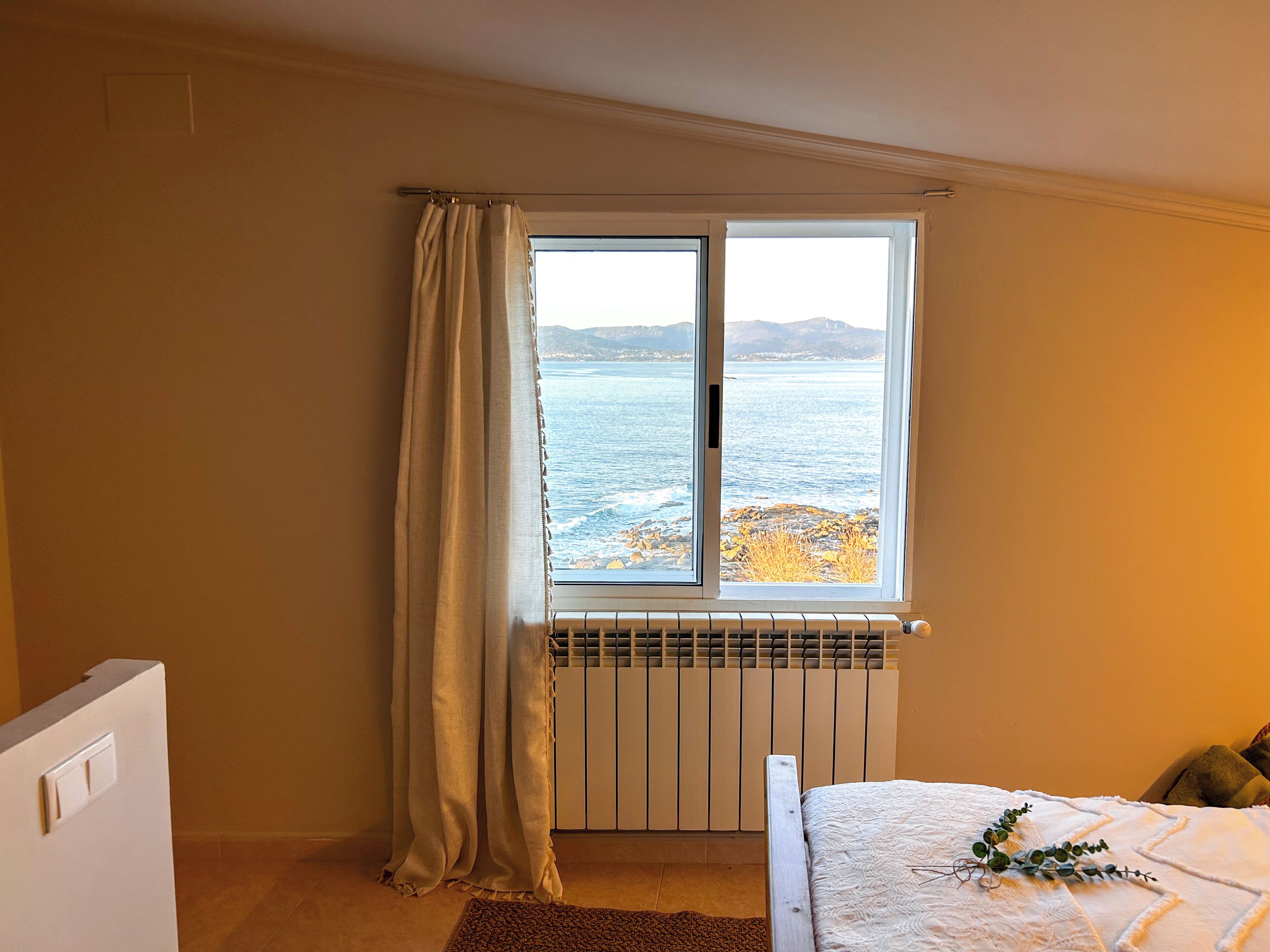 Ein Schlafzimmer mit Meerblick durch ein Fenster, einem Heizkörper, einem Bett mit Decke und einer Pflanze.