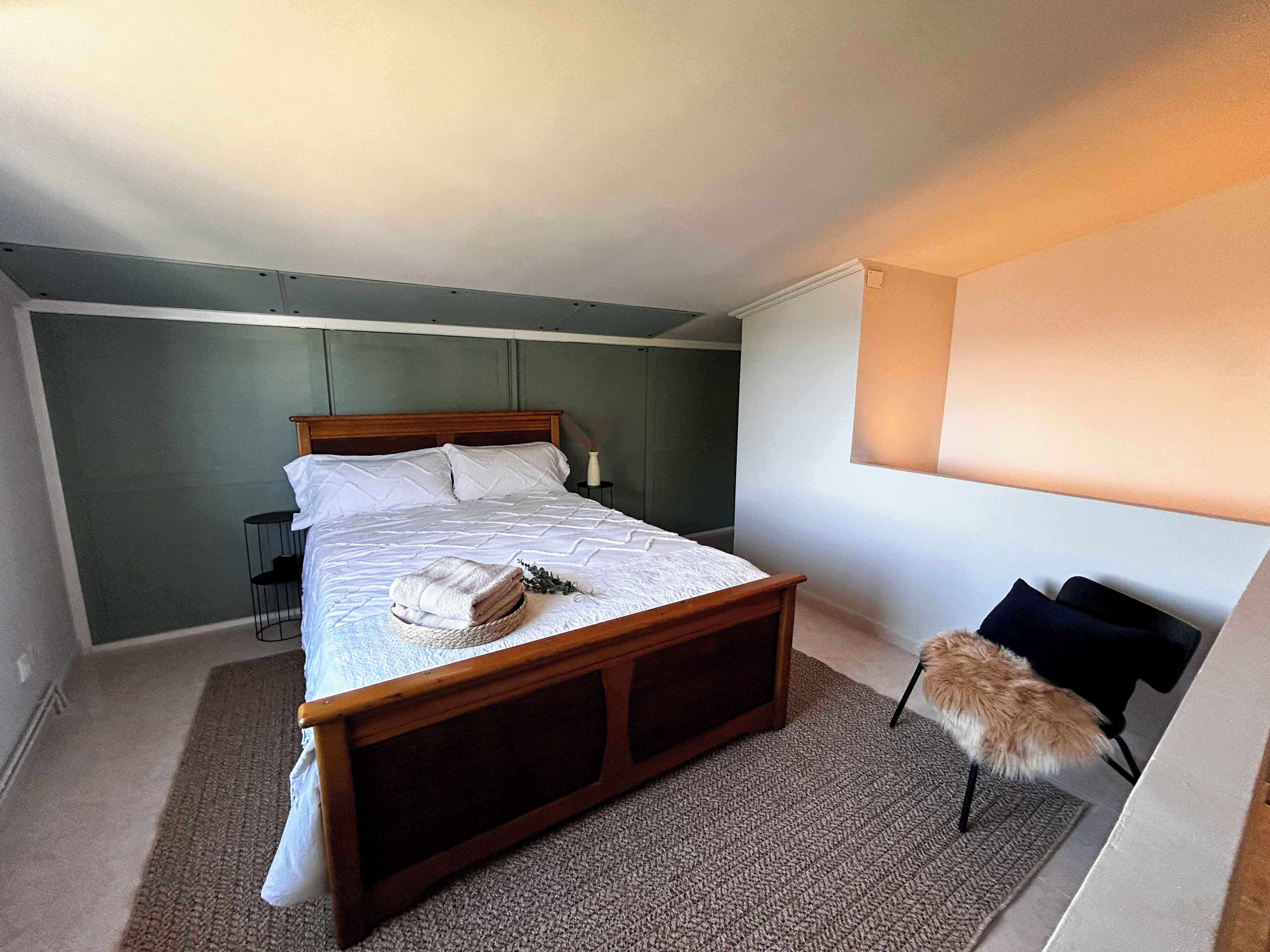 A neatly made bed with a wooden headboard, a small bedside table with a lamp, and a chair with a fur throw.