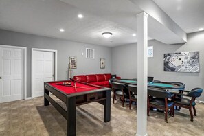 Una habitación con mesa de billar, un sofá de cuero rojo, una mesa de cartas y sillas.