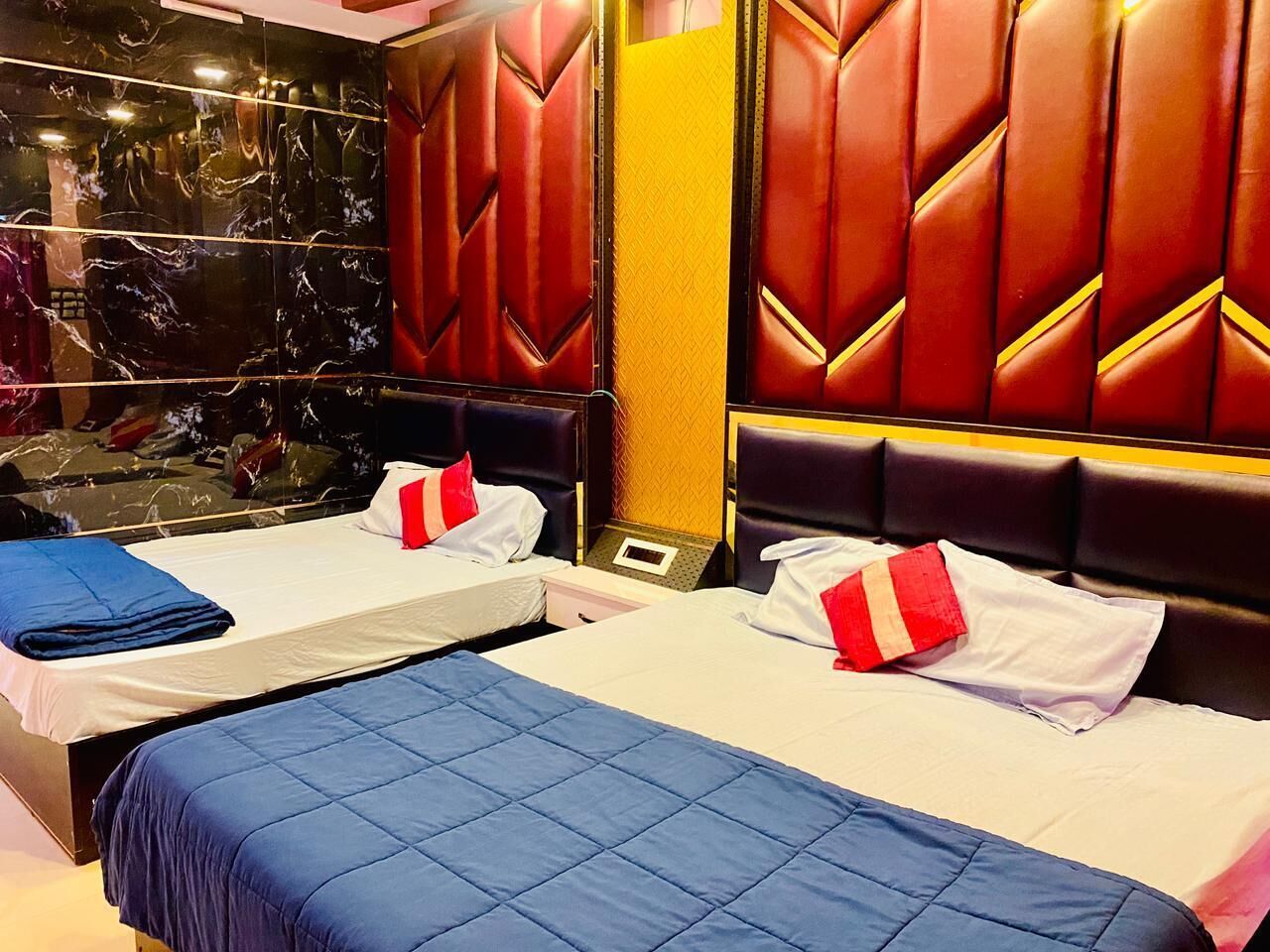 A hotel room with two beds, each with a blue quilt and red and white pillows. The room features a marble wall and a red leather headboard.
