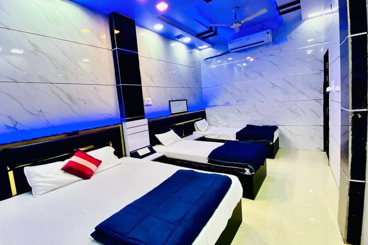 Une chambre d’hôtel avec trois lits, chacun agrémenté de draps blancs et de couvertures bleues. Les murs sont recouverts de carreaux de marbre blanc, et un ventilateur de plafond est visible.