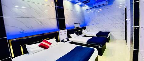 A hotel room with three beds, each with white linens and blue bedding. The walls are covered in white marble tiles, and there is a ceiling fan visible.