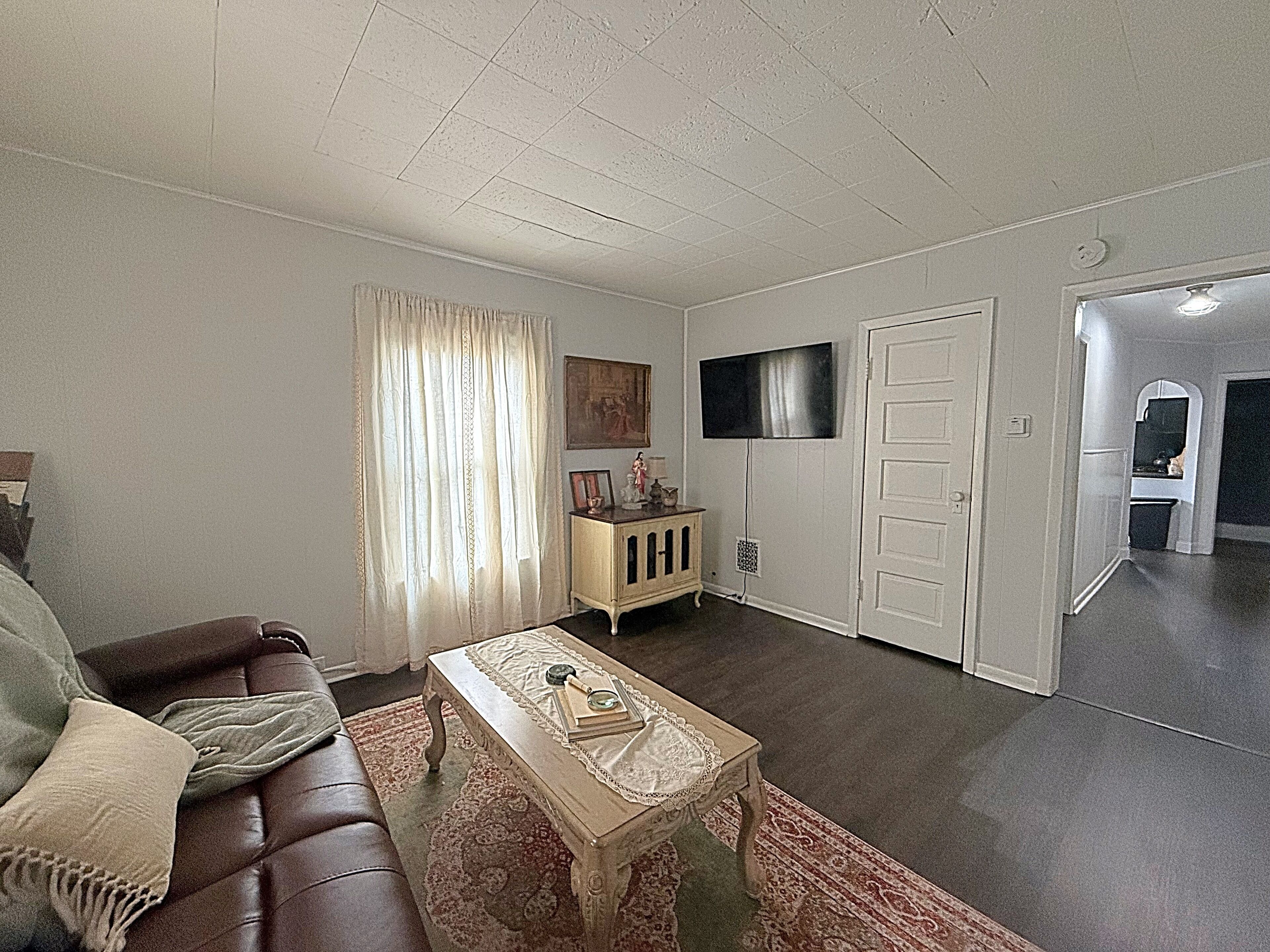 Ein Wohnzimmer mit einem braunen Ledersofa, einem Couchtisch mit Büchern und einer Lupe, einer kleinen Vitrine und einem Fernseher an der Wand.
