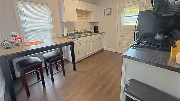 A compact kitchen with white cabinets, a black countertop, and a gas stove. There are two bar stools and a window with blinds.