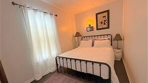 A bedroom with a bed, white curtains, a bedside lamp, a nightstand, and a framed picture on the wall.