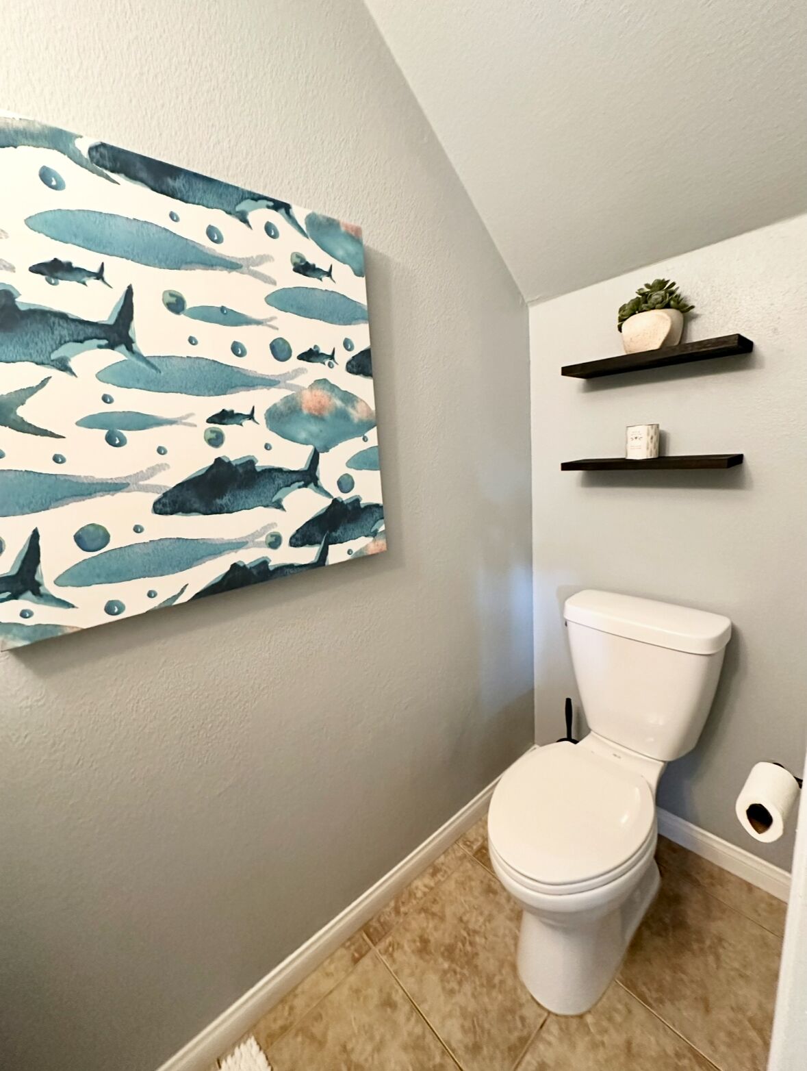 A bathroom with a white toilet, a blue and white abstract painting, and two dark wooden shelves with a plant and a candle.