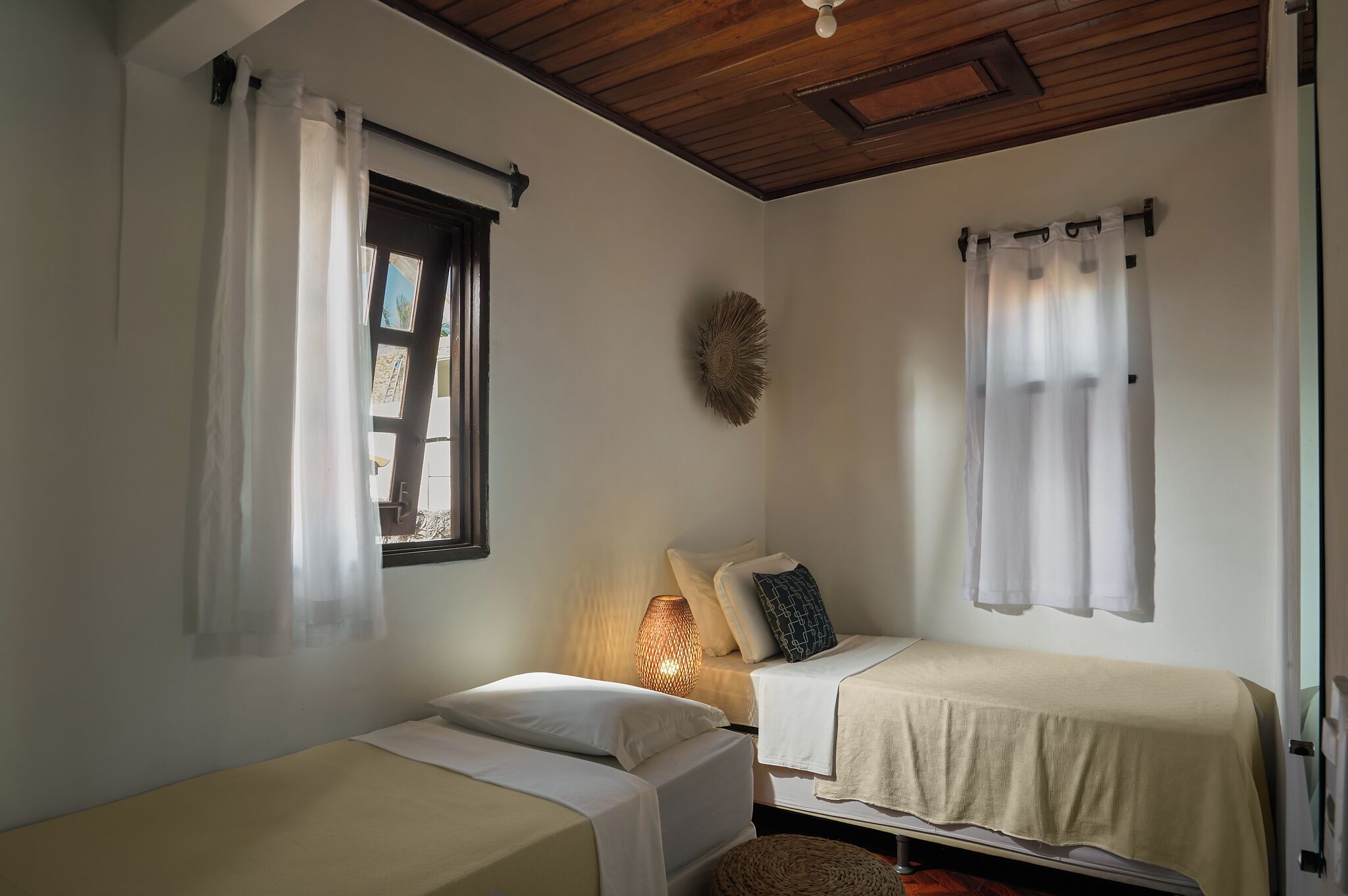 A small, cozy room with two single beds, a window with curtains, a wall-mounted lamp, and a decorative wall piece.