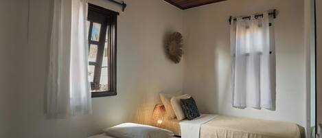 A small, cozy room with two single beds, a window with curtains, a wall-mounted lamp, and a decorative wall piece.