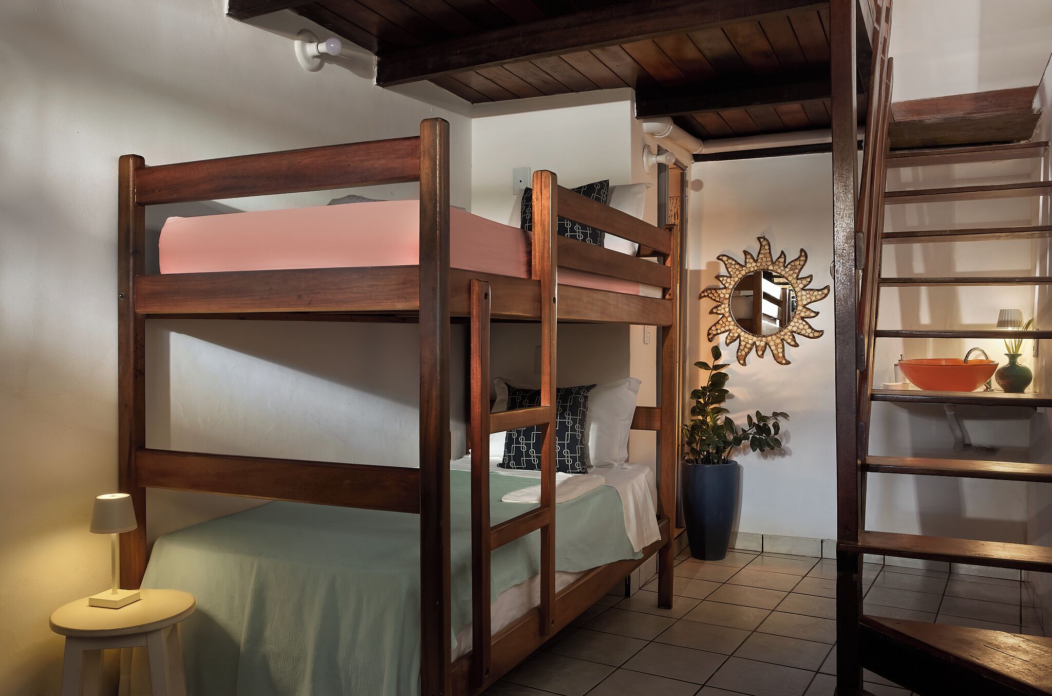 A bunk bed room with a wooden frame, a small table with a lamp, a potted plant, and a shelf with a mirror and decorative items.