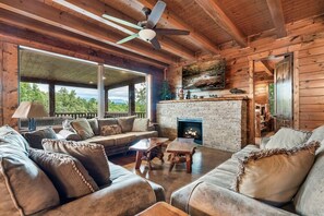 A log cabin interior with a stone fireplace, wooden furniture, and a large window overlooking greenery. - Indoor Golf Cabin w/ Amazing Views! Sleeps 40! (Sevierville)