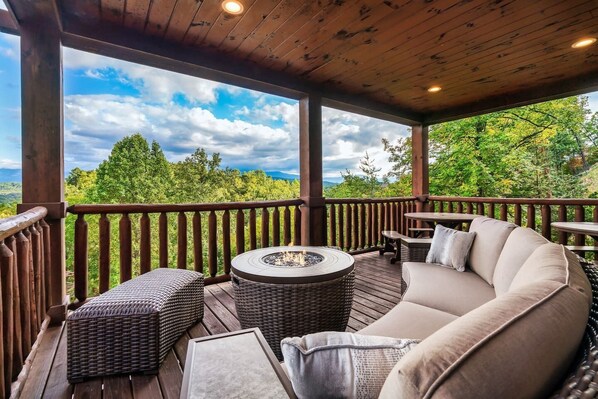 A covered outdoor deck with a fire pit, wicker furniture, and a cushioned seating area.