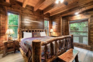 Une chambre dans un chalet en rondins, avec un lit en bois, deux lampes de chevet, une fenêtre donnant sur la verdure et une tête de lit en bois.
