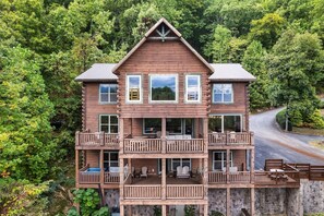 A two-story log cabin with multiple balconies, a stone foundation, and a surrounding forest.