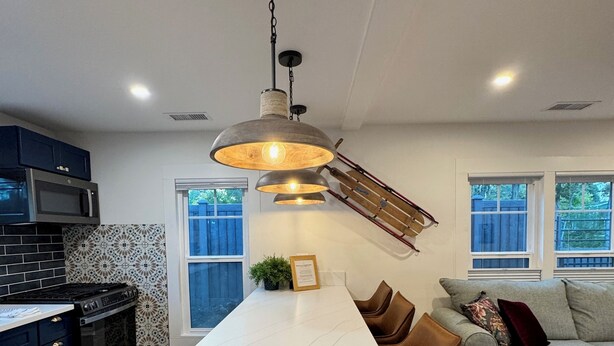 Una cocina moderna con una isla central, armarios oscuros, un microondas y una estufa. Hay una zona de comedor con sillas marrones y un sofá.