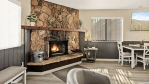 Un salon chaleureux avec une cheminée en pierre, un coin repas avec une table et des chaises, et une fenêtre avec des stores.