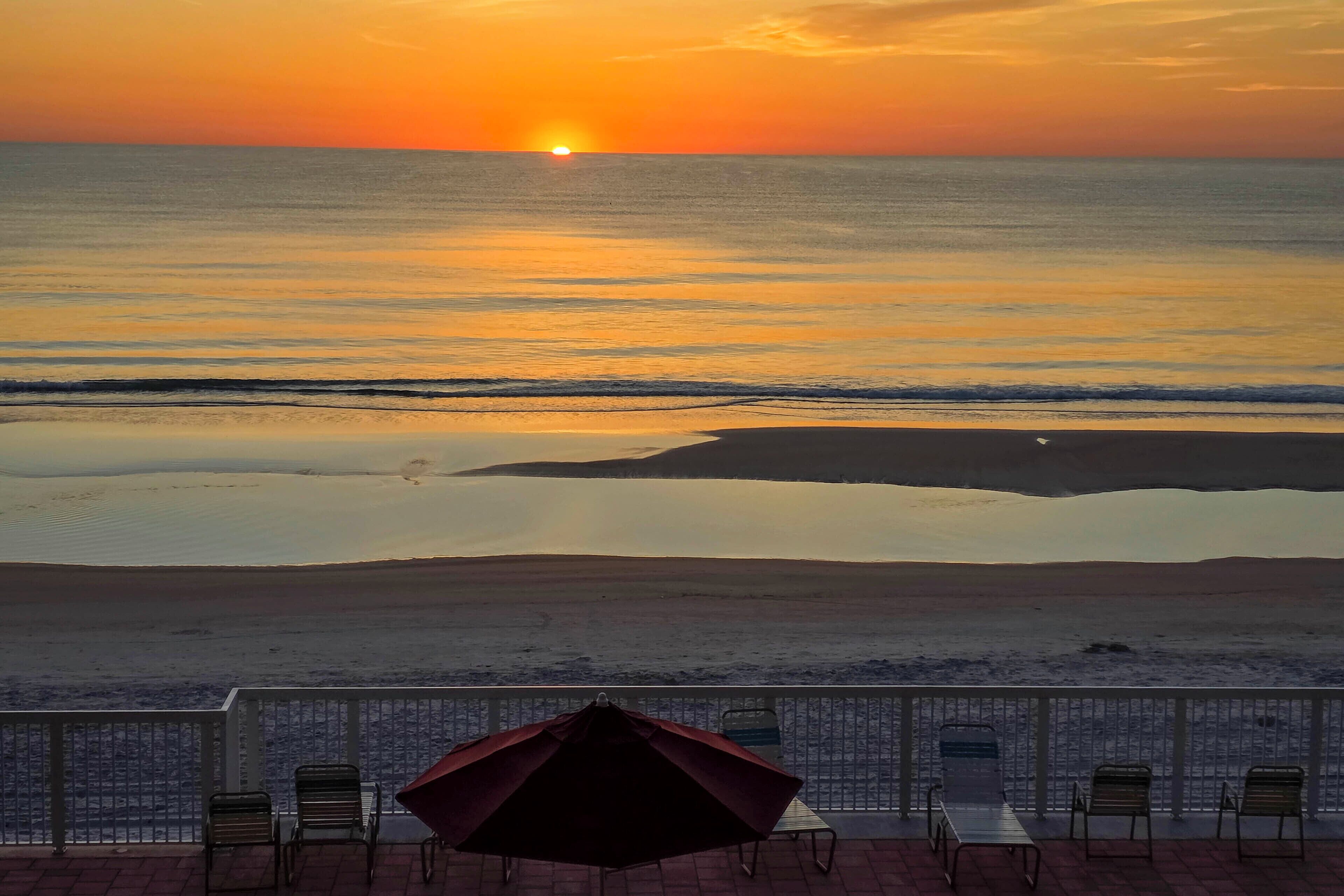 Vista a la playa con atardecer, sillas y una sombrilla en una terraza.