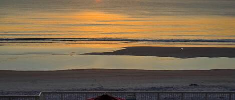 Vista a la playa con atardecer, sillas y una sombrilla en una terraza.
