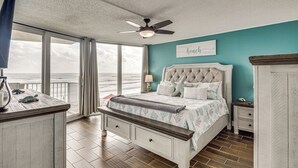 A bedroom with a large window overlooking the ocean, a ceiling fan, a bed with a tufted headboard, and a nightstand with a lamp. - Pool, Balcony & More! Oceanfront Shores Club Condo (Daytona Beach Shores)