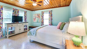 Un dormitorio con cama, televisor sobre un tocador, ventilador de techo y ventana con cortinas.