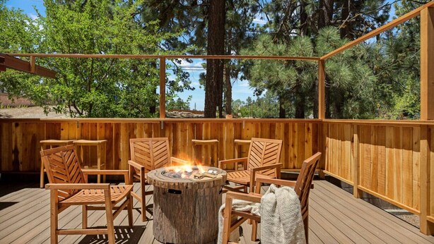 A wooden deck with a fire pit surrounded by wooden chairs and tables, set against a backdrop of trees and a clear sky. - Lawliss Hideout, Steps to Village w/ Hot Tub (Big Bear Lake)