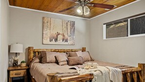 A bedroom with a wooden bed frame, a ceiling fan, a painting of bears, and a window with a view. - Lawliss Hideout, Steps to Village w/ Hot Tub (Big Bear Lake)