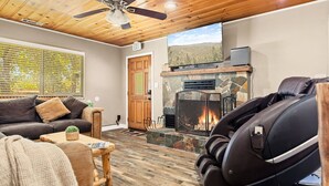 A cozy living room with a fireplace, comfortable seating, and a ceiling fan. - Lawliss Hideout, Steps to Village w/ Hot Tub (Big Bear Lake)