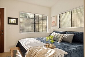 Ein Schlafzimmer mit einem Bett, zwei Fenstern mit Jalousien, einem Nachttisch mit Lampe und einem gerahmten Bild an der Wand.