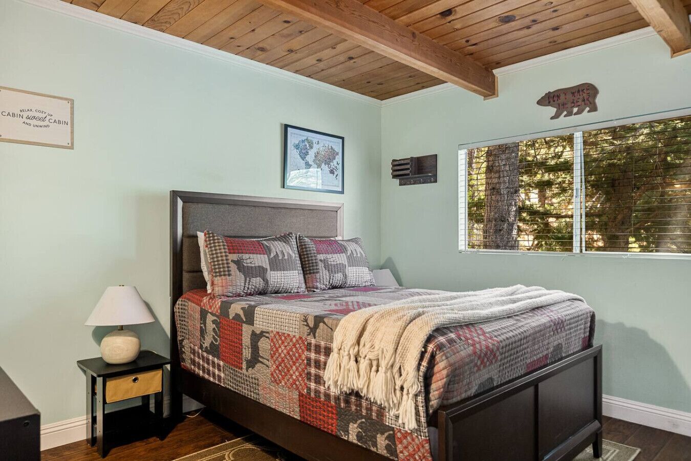 Un dormitorio con cama, mesita de noche, lámpara, un cuadro enmarcado, ventana con vista a árboles y un cartel con forma de oso en la pared.