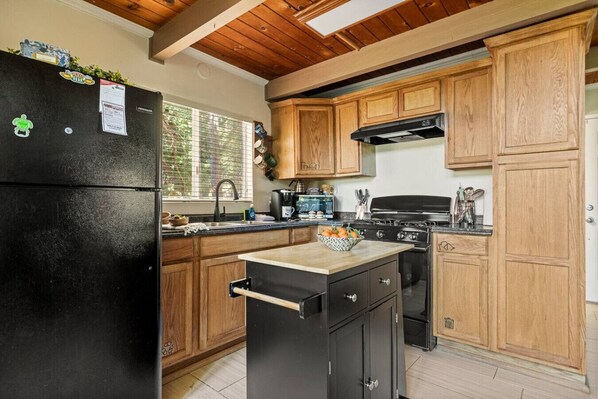 A kitchen with wooden cabinets, a black refrigerator, a stove, and a small island with a bowl of fruit.