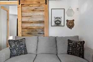 A grey sofa with patterned cushions, a wooden wall panel, a framed raccoon picture, and a bear head decoration.