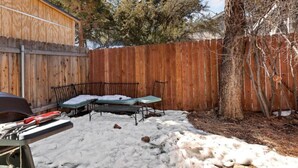 A backyard with a wooden fence, a metal table, and a bench covered in snow. - Modern Chic Spruce Retreat - Hot Tub + AC + Pets (Big Bear)