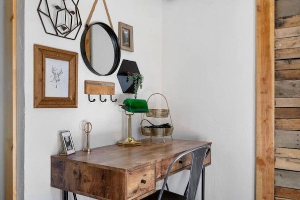 Un bureau en bois, une lampe verte, une chaise en métal et des éléments de décoration fixés au mur, notamment un miroir, des tableaux encadrés et un luminaire au design géométrique.