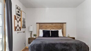 A bedroom with a wooden headboard, a bed with a grey blanket, two pillows, a nightstand with a lamp, and a wall with a framed picture and a sign. - Modern Chic Spruce Retreat - Hot Tub + AC + Pets (Big Bear)