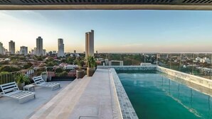 Una piscina en la azotea con vistas a la ciudad, sillas de descanso y plantas en macetas.