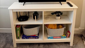 Eine weiße Unterhaltungsanlage mit einem Flachbildfernseher, einer Fernbedienung, einem Router und Regalen mit Gesellschaftsspielen und einem Korb.