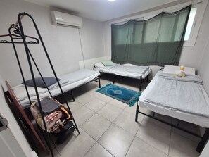 Un dormitorio con dos camas individuales, una mesita con silla, un radiador portátil, una ventana con cortina y un aire acondicionado de pared.