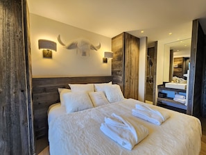 Quarto de hotel com uma cama grande, duas luminárias fixadas na parede, cabeceira de madeira e um espelho refletindo o banheiro.